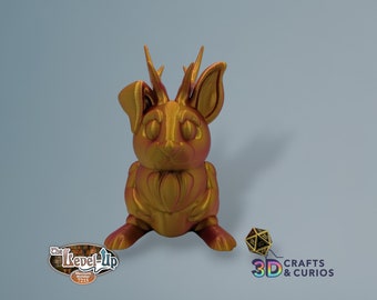 May include: A gold-colored 3D printed rabbit figurine with antlers. The rabbit is sitting on a light blue surface. The rabbit has a detailed design and a textured surface.