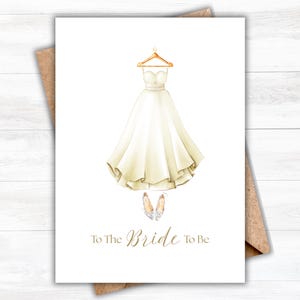 Puede incluir: Una tarjeta de felicitación con una ilustración en acuarela de un vestido de novia en una percha, zapatos de novia y el texto "To The Bride To Be" en letras doradas. La tarjeta está sobre un fondo blanco con un sobre marrón.