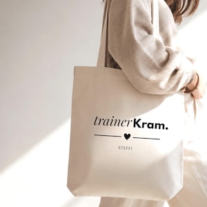 Puede incluir: Un bolso de tela de color crema con el texto negro impreso trainerKram. sobre una línea horizontal con un pequeño icono de corazón en el centro y el nombre STEFFI impreso debajo de la línea. El bolso tiene dos correas.