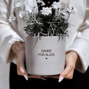 Peut inclure: Un pot de fleurs cylindrique gris avec des fleurs blanches et du feuillage vert. Le pot porte l'inscription "DANKE FÜR ALLES" et un petit symbole de cœur. Le pot est tenu par une personne portant un pull blanc.
