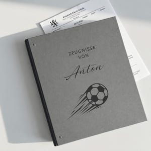 Puede incluir: Una carpeta gris con un remache metálico, que presenta el texto impreso ZEUGNISSE VON Anton y un gráfico negro de un balón de fútbol en movimiento. Un documento blanco de la Friedrich-Ebert-Schule es parcialmente visible detrás.