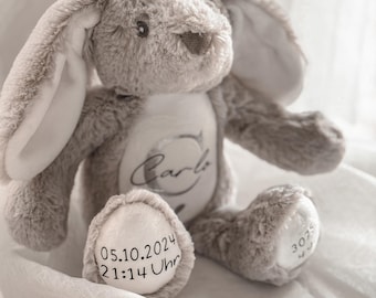 May include: A plush gray bunny rabbit with long ears and a white belly. The bunny has the name "Carlo" embroidered on its belly. The date "05.10.2024 21:14 Uhr" is embroidered on one foot. A soft toy for a baby.