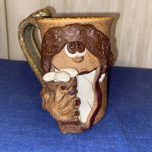 Può includere: Una tazza in ceramica stravagante con un design a volto. La tazza presenta un esterno marrone strutturato con un volto scolpito, inclusi capelli ricci, occhi grandi e una bocca sorridente. Il manico è un design curvo e strutturato. L'interno è bianco.