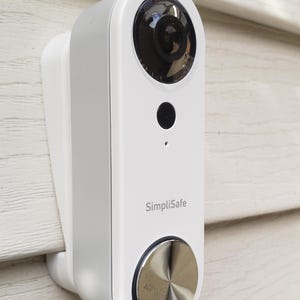 Puede incluir: Una cámara de timbre SimpliSafe blanca montada en una pared de revestimiento blanco. La cámara tiene una lente gran angular y un botón plateado.