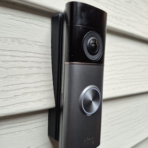 May include: A black and grey Ring video doorbell fixed to a white siding wall. The doorbell features a camera lens, a silver button, and the Ring logo. It is designed for home security and surveillance purposes.