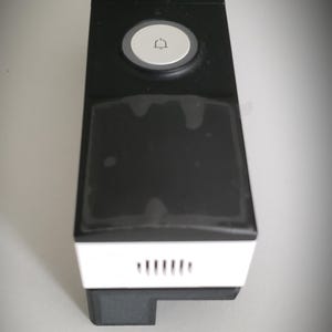 May include: A black and white rectangular doorbell with a white button featuring a bell icon. The doorbell has a white bottom section with ventilation slits and a sticker on top.
