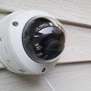 May include: A white security camera mounted on a white siding wall. The camera has a clear dome lens and a black interior.