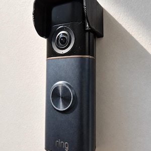 May include: A black and grey smart doorbell with a built-in camera and a silver button. The doorbell has a protective black hood and is mounted on a white wall. The brand name "ring" is visible.