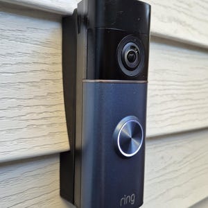 Op de afbeelding: Een zwarte en grijze Ring videodeurbel gemonteerd op een lichtbeige gevelbekleding. De deurbel heeft een cameralens, een zilveren knop en het Ring-logo. Het apparaat is ontworpen voor huisbeveiliging en smart home integratie.