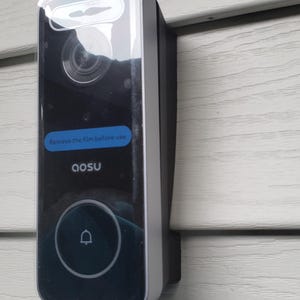 May include: A black and silver doorbell camera with a clear protective film over the lens. The camera has a button with a bell icon and the text "Remove the film before use" and "AOSU" on the screen.