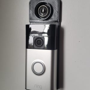 May include: A silver and black Ring video doorbell mounted on a wall. The doorbell has a camera, a button, and the Ring logo. Above the doorbell is a black camera mount.