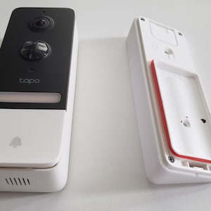 Puede incluir: Un sistema de timbre inteligente blanco y negro con cámara, botón y altavoz. El timbre tiene la marca "tapo". El soporte de montaje es blanco con un sello de goma rojo.