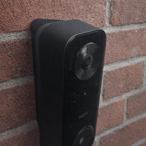May include: A black Aqara video doorbell mounted on a red brick wall. The doorbell has a camera lens and a speaker. The Aqara logo is visible on the device.