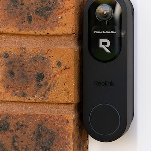 May include: A black Reolink video doorbell mounted on a white surface next to a brick wall. The doorbell has a camera lens, the text "Please Before Use", the letter "R", and the brand name "Reolink".