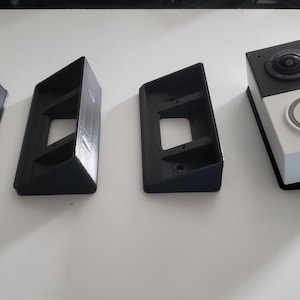 May include: A smart doorbell in black and white, featuring a camera, a button, and a speaker. Three black angled mounting brackets are also present, designed to hold the doorbell. The items are on a white surface.