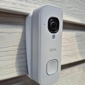Può includere: Un videocitofono Blink bianco con corpo rettangolare, obiettivo della fotocamera nero nella parte superiore, una piccola spia luminosa e un pulsante quadrato nella parte inferiore. Il dispositivo è montato verticalmente su un rivestimento orizzontale chiaro.