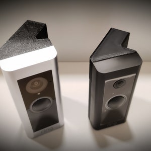 May include: Two security cameras are shown, one silver and one black. Each camera has a rectangular body with a lens and speaker. The top of each camera has a textured, angled cover. These cameras are designed for home security.