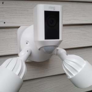 May include: A white Ring security camera with a motion sensor and two white floodlights mounted on a white siding wall. The camera has a black lens and the text "ring" is printed on the front of the camera.