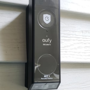 Peut inclure: Une sonnette Eufy Security noire avec un logo de bouclier blanc et le texte "Eufy Security" et "PET 1 Raccolta Plastica".