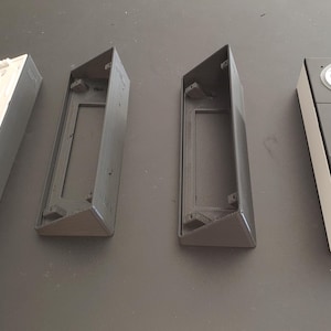 May include: Four white and grey plastic housings for a doorbell camera system. The housings are rectangular and have a slot for a camera lens. The doorbell camera system has a white faceplate with two buttons and a black camera lens.