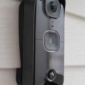 May include: A black Swann security camera mounted on a white wall. The camera has a wide-angle lens, a doorbell button, and a motion sensor.