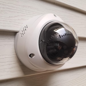 May include: A white dome security camera with a clear, rounded lens is mounted on a light-colored, textured wall. The camera has a sleek design with ventilation slots and a small screw. The lens reflects the surrounding environment.