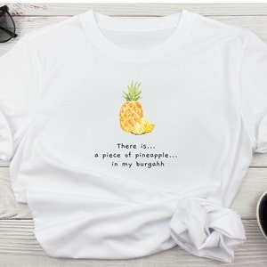 May include: White t-shirt with a graphic of a pineapple and the text "There is... a piece of pineapple... in my burgahh."