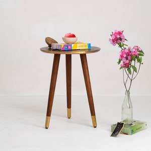 May include: A small, round side table with a brass-colored top and three wooden legs. The legs have gold-colored tips. On the table are a small bowl with a red apple, a wooden bowl, and a book. A vase with pink flowers is to the side.