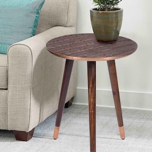 May include: A round, brown side table with a patterned top and three dark wooden legs. The legs have copper-colored tips. A potted plant sits on the table. The table is next to a beige sofa with a blue pillow.