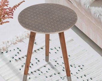 Op de afbeelding: Een kleine, ronde bijzettafel met een patroon op het blad en drie houten poten. Het tafelblad heeft een herhalend geometrisch ontwerp in bruin- en beigetinten. De poten zijn van natuurlijk hout met zilverkleurige uiteinden.