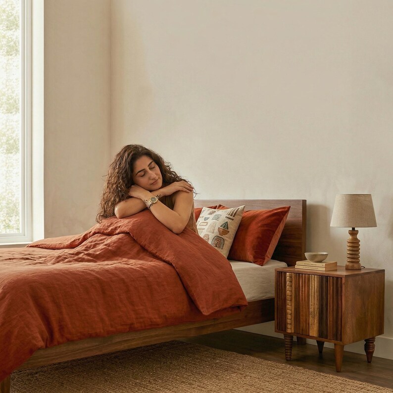 Pu&ograve; includere: Scena di camera da letto con un letto coperto da un piumino e cuscini color ruggine. Un comodino in legno con una lampada e oggetti decorativi &egrave; accanto al letto. La stanza ha una palette di colori neutri.