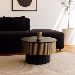 May include: A modern, two-tiered coffee table with a black top and base. The middle section features vertical gold-colored slats. A small green and gold-rimmed vase sits on top. The table is on a beige rug in a living room setting.