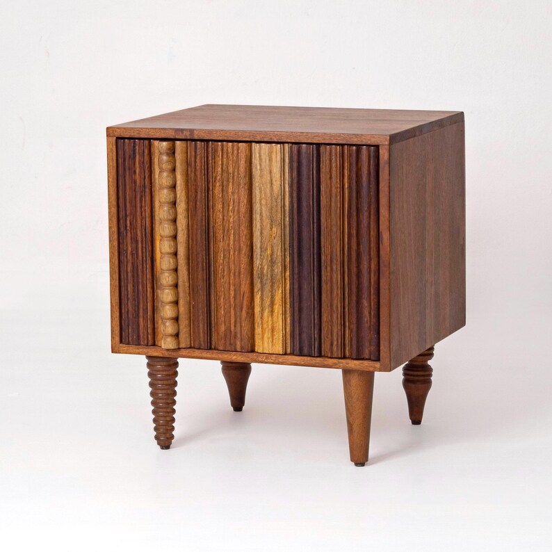 Pu&ograve; includere: Un comodino in legno in stile moderno della met&agrave; del secolo, di forma rettangolare. La parte anteriore presenta doghe verticali in legno in varie tonalit&agrave; di marrone e beige. Ha quattro gambe affusolate, una con un design a spirale.