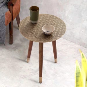 May include: A small, round side table with a patterned gold top and three wooden legs. The legs have gold-coloured tips. A green cup and a small bowl with blue floral designs sit on the table. The table is approximately 50 cm in diameter.