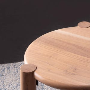 May include: A round wooden stool featuring a smooth, light-toned circular top and three thick, cylindrical legs that connect to the surface with visible circular joints. The natural wood grain is clearly visible across the entire structure.