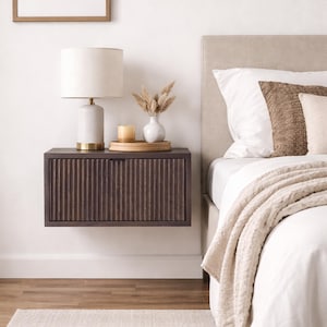 Può includere: Un comodino marrone scuro, montato a parete, con un design a scanalature verticali. Una lampada bianca con base dorata, un piccolo vaso bianco con fiori secchi e una candela sono appoggiati sopra. Il comodino è accanto a un letto con lenzuola bianche e una testiera beige.