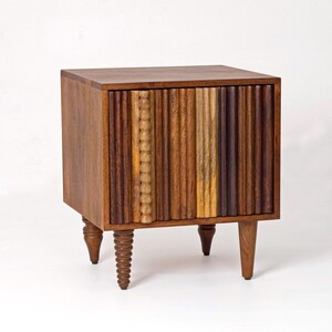 Pu&ograve; includere: Un comodino in legno con un fronte a coste verticali multicolore. Il comodino ha un piano quadrato ed &egrave; sostenuto da quattro gambe corte e tornite. Le tonalit&agrave; del legno variano dal marrone chiaro allo scuro.