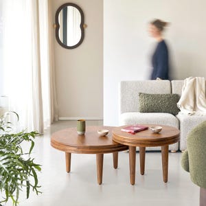 Könnte beinhalten: Zwei runde Couchtische aus Holz mit konisch zulaufenden Beinen, die ineinander verschachtelt sind. Die Tische haben eine warme braune Holzmaserung. Eine kleine grüne Keramiktasse und eine weiße Schale stehen auf dem größeren Tisch.