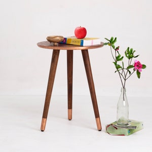 May include: A small, round side table with a copper-colored top and three wooden legs. The legs have copper-colored tips. A red apple, a small wooden bowl, and a book are on the table. A vase with flowers is next to the table.