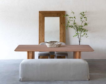 May include: A modern dining room setup featuring a rectangular wooden table with a light brown finish, supported by thick, cylindrical legs. A light gray bench sits in front of the table. Two gray chairs are behind the table. A large wooden framed mirror is in the background.