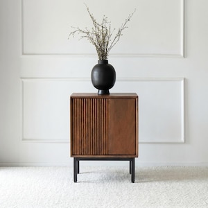 May include: A wooden cabinet with a ribbed front panel and a smooth panel on the right side. The cabinet is supported by black metal legs. A black vase with dried branches sits on top of the cabinet.