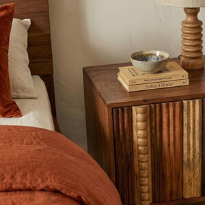 Pu&ograve; includere: Un comodino in legno con un fronte intagliato, con libri e una piccola ciotola. I libri recitano "Mid Century Design Principles" e "Natural Home". Una lampada con base in legno &egrave; sul comodino. Il letto ha lenzuola color ruggine e un cuscino di velluto.