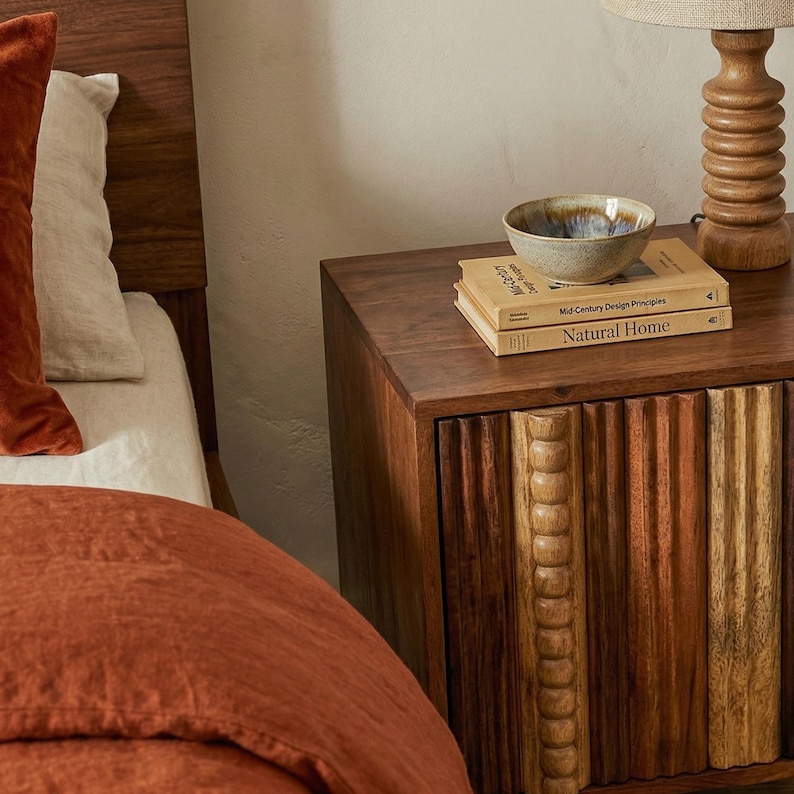 Pu&ograve; includere: Un comodino in legno con un fronte intagliato, con libri e una piccola ciotola. I libri recitano "Mid Century Design Principles" e "Natural Home". Una lampada con base in legno &egrave; sul comodino. Il letto ha lenzuola color ruggine e un cuscino di velluto.
