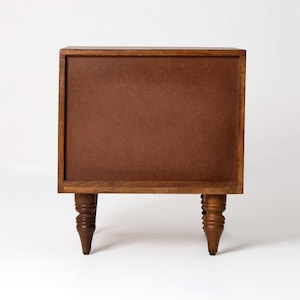 Pu&ograve; includere: Un piccolo comodino in legno con finitura marrone scuro. Il comodino ha una forma rettangolare con un pannello marrone al centro e quattro gambe tornite. Le gambe sono alte circa 15 cm.
