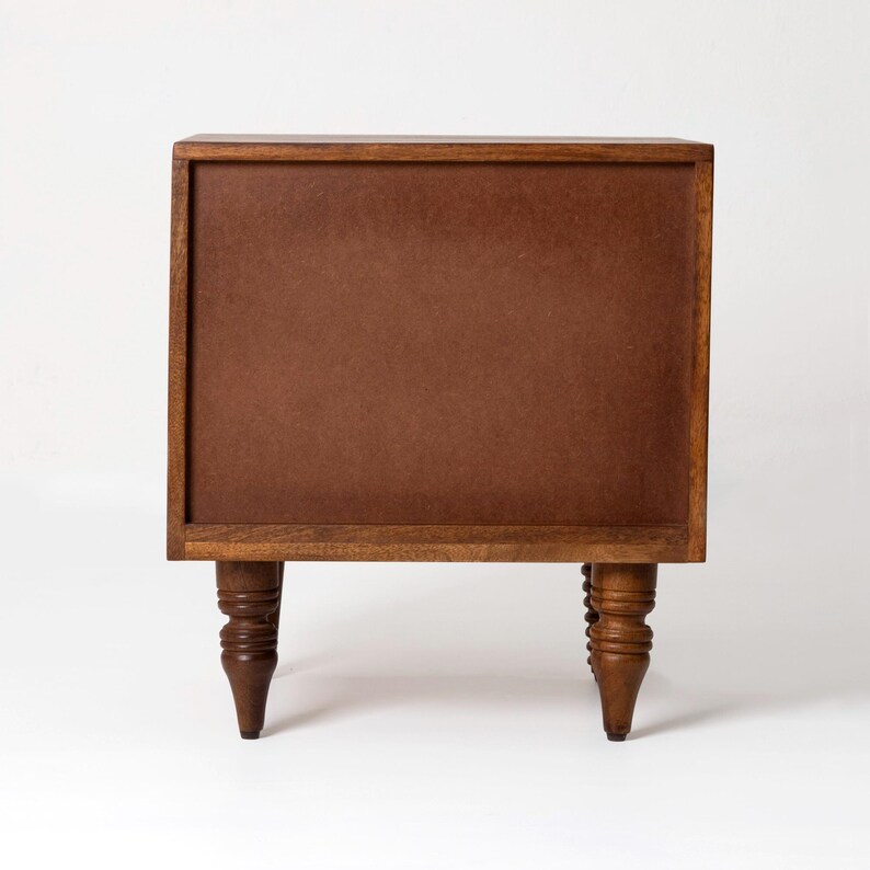Pu&ograve; includere: Un piccolo comodino in legno con finitura marrone scuro. Il comodino ha una forma rettangolare con un pannello marrone al centro e quattro gambe tornite. Le gambe sono alte circa 15 cm.