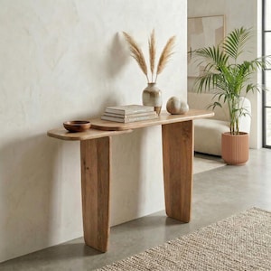 May include: A light brown wooden console table with an asymmetrical design. The table has a curved top with a small, attached shelf. A small wooden bowl, books, and decorative items sit on the table. The table is 152 cm long.