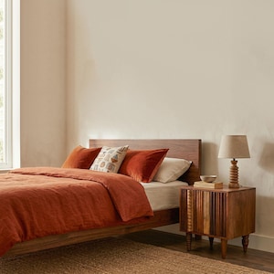 Pu&ograve; includere: Una scena di camera da letto con un letto in legno, biancheria da letto e cuscini color ruggine. Un cuscino fantasia aggiunge un tocco decorativo. Un comodino in legno con una lampada e una piccola ciotola si trova accanto al letto. La stanza ha una palette di colori neutri.