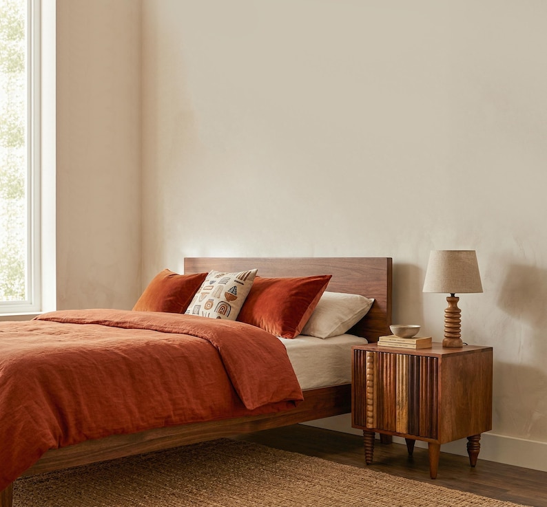 Pu&ograve; includere: Una scena di camera da letto con un letto in legno, biancheria da letto e cuscini color ruggine. Un cuscino fantasia aggiunge un tocco decorativo. Un comodino in legno con una lampada e una piccola ciotola si trova accanto al letto. La stanza ha una palette di colori neutri.