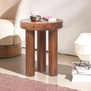 May include: A round, dark brown wooden side table with three cylindrical legs. The tabletop holds a small decorative bowl and a book. The table is a stylish accent piece for a living room or bedroom.