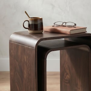 Op de afbeelding: Een donkerbruine houten bijzettafel met een gebogen ontwerp. Een keramische mok met een lepel, een boek en een bril liggen op de tafel. De mok heeft bruine en bruinachtige tinten. Het boek is lichtbruin.